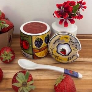 Peut inclure: Un pot à confiture en céramique avec un couvercle, rempli de confiture rouge. Le pot est décoré d'images de cerises, de mûres et d'une tranche de citron. Une cuillère blanche et des fraises fraîches sont également présentes.