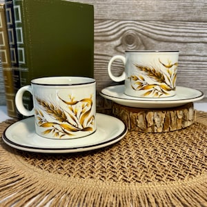 May include: Two vintage cream-colored coffee cups and saucers with a wheat design in shades of gold and brown. The cups have a black rim and handle, and the saucers have a matching black border. The set is displayed on a woven placemat.