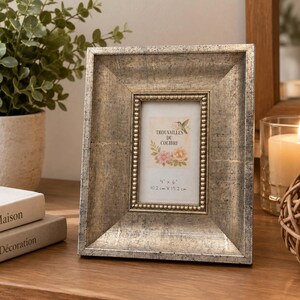 Peut inclure: Un cadre photo rectangulaire avec une finition argentée vieillie et une bordure intérieure perlée. Le cadre contient une carte avec une illustration de colibri, des fleurs et le texte "Trouvailles du Colibri" avec "4 x 6" et "10,2 cm x 15,2 cm" imprimés en dessous.