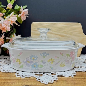 Op de afbeelding: Een witte ovenschaal met een doorzichtig glazen deksel. De schaal is versierd met een bloemenpatroon in pastelkleuren, waaronder blauw, roze en geel. De schaal heeft twee handvatten en staat op een wit kanten kleedje.