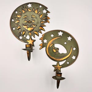 May include: Two celestial-themed wall sconces. One is a smiling sun with star cutouts, the other a crescent moon with stars. Both have a green and gold finish and candle holders. Decorative home decor.