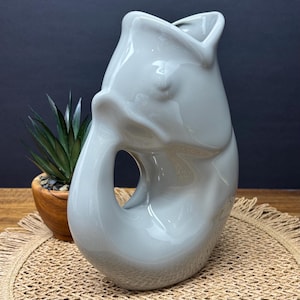 May include: A light grey ceramic vase with an unusual, abstract design. The vase has a curved, organic shape with a large opening and a smooth, glossy finish. The vase is displayed on a wooden surface with a small plant in a wooden pot.