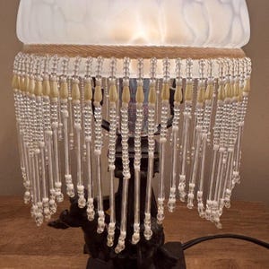 K&ouml;nnte beinhalten: Eine Tischlampe mit einem mattwei&szlig;en Glasschirm und einem dekorativen Behang aus klaren Glasperlen sowie cremefarbenen tropfenf&ouml;rmigen Akzenten. Der dunkle Lampenfu&szlig; ist mit Cherub-Figuren verziert und tr&auml;gt den beleuchteten Glasschirm.