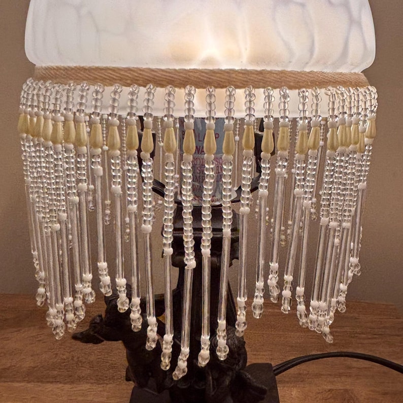 K&ouml;nnte beinhalten: Eine Tischlampe mit einem mattwei&szlig;en Glasschirm und einem dekorativen Behang aus klaren Glasperlen sowie cremefarbenen tropfenf&ouml;rmigen Akzenten. Der dunkle Lampenfu&szlig; ist mit Cherub-Figuren verziert und tr&auml;gt den beleuchteten Glasschirm.