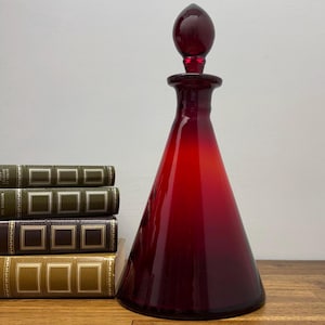 Może przedstawiać: Rubinowa szklana karafka o stożkowym kształcie i pasującym korku. Obok stos zabytkowych książek ze złotymi literami i dekoracyjnymi okładkami. Karafka to przedmiot dekoracyjny.