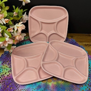 May include: Three pink divided plates. Each plate has a square shape with rounded corners and a central divider, creating four separate compartments. The plates are arranged on a colourful, patterned surface, with flowers in the background.