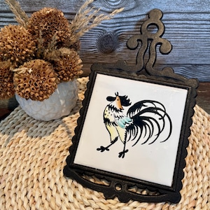 Pode incluir: Um descanso de panela de metal preto com um azulejo decorativo de galo. O azulejo apresenta uma ilustração colorida de um galo sobre um fundo branco. O descanso de panela tem uma borda ondulada e uma alça. Um vaso branco com flores secas está no fundo.