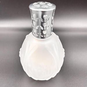 Puede incluir: Una lámpara de fragancia de vidrio esmerilado con un cuerpo redondeado y texturizado que presenta patrones florales en relieve. La parte superior es una tapa de metal pulido color plata con un diseño recortado de tulipanes y corazones.