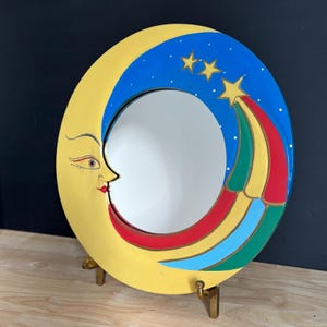 May include: A whimsical, oval-shaped mirror featuring a yellow crescent moon with a smiling face. The top portion is blue with white stars. A colorful rainbow of red, yellow, green, and blue emanates from three gold stars. The mirror is supported by a gold stand.