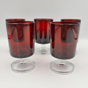Pode incluir: Um conjunto de cinco copos com taças em vermelho profundo e hastes e bases circulares em vidro transparente. Cada copo tem uma forma cilíndrica lisa com uma borda arredondada, exibindo um design consistente em todas as cinco peças.