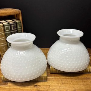 Könnte beinhalten: Zwei weiße Lampenschirme aus Hobnail-Glas mit runder Form und ausgestelltem Oberteil. Die Schirme stehen auf alten Büchern. Der Hintergrund ist dunkel.