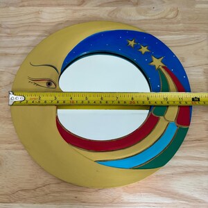 May include: A decorative, round mirror with a crescent moon design. The moon is painted gold with a detailed eye. The top portion features a blue sky with three stars and a colorful shooting star. The mirror measures approximately 11 inches in diameter.