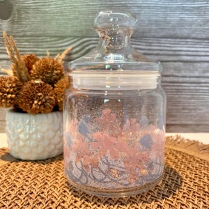 Puede incluir: Un tarro de cristal transparente con tapa, decorado con diseños florales rosas y azules. El tarro está lleno de purpurina y tiene un pomo decorativo. El tarro está sobre una esterilla tejida.