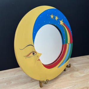 May include: A whimsical, round mirror in the shape of a crescent moon. The moon is painted yellow with a smiling face. The top of the moon is blue with three gold stars and a colorful shooting star design. The mirror is supported by a gold-colored stand.