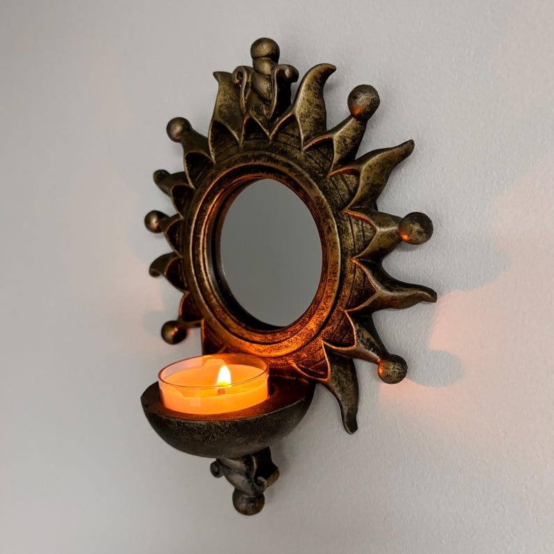 May include: A wall-mounted candle holder featuring a circular mirror surrounded by a sunburst frame with a dark bronze finish. A small, clear glass tea light holder is attached at the bottom, containing a lit candle with a warm orange glow.