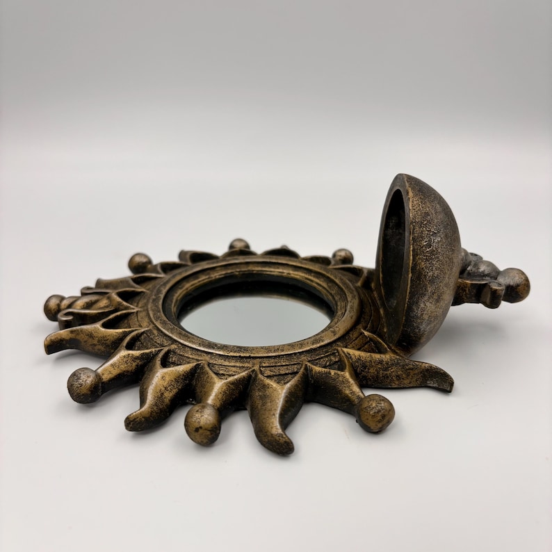 May include: A circular, sun-shaped wall mirror with a distressed bronze finish. The frame features radiating rays tipped with small spheres, surrounding a central reflective glass surface. A hinged, dome-shaped cover is attached to the side.