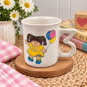 Peut inclure: Un mug en céramique blanche orné d'une illustration de poupée aux couettes brunes, vêtue d'une robe jaune et tenant trois ballons colorés. La tasse possède une anse en forme de lettre B, offrant une silhouette originale à cet objet.