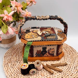 May include: A hexagonal wooden sewing box featuring a cat tapestry pattern. It has a handle and button closure. A green tape measure, spools of thread, and wooden pegs are placed nearby. The box rests on a woven mat.