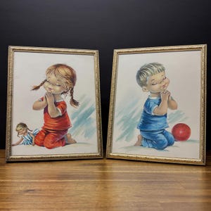 Könnte beinhalten: Zwei gerahmte Vintage-Drucke. Einer zeigt ein Mädchen in rotem Schlafanzug mit Zöpfen, das betend kniet, und eine Puppe. Der andere zeigt einen Jungen in blauem Schlafanzug, der mit einem roten Ball kniet. Beide haben goldene Rahmen.