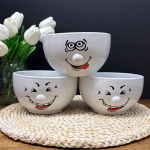 Pode incluir: Três tigelas de cerâmica branca empilhadas, cada uma com um desenho de rosto sorridente. Os rostos têm olhos pretos, sobrancelhas e sorrisos com línguas vermelhas. As tigelas estão sobre um tapete de mesa tecido, com flores brancas em um vaso ao fundo.