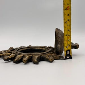 May include: A bronze-colored sun-shaped wall sconce with a textured finish. A yellow measuring tape is positioned vertically against the side, showing the sconce has a height of approximately 4 inches from the base to the top edge.