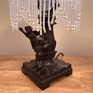 K&ouml;nnte beinhalten: Eine Tischlampe mit einem dunkelbraunen Sockel, der zwei skulptierte Puttenfiguren zeigt. Der Lampenschirm ist mit zahlreichen herabh&auml;ngenden Str&auml;ngen aus klaren und wei&szlig;en Perlen verziert, die einen Frangeneffekt um die Struktur erzeugen.