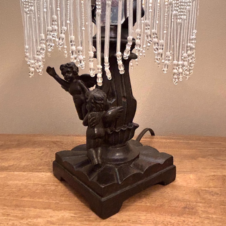 K&ouml;nnte beinhalten: Eine Tischlampe mit einem dunkelbraunen Sockel, der zwei skulptierte Puttenfiguren zeigt. Der Lampenschirm ist mit zahlreichen herabh&auml;ngenden Str&auml;ngen aus klaren und wei&szlig;en Perlen verziert, die einen Frangeneffekt um die Struktur erzeugen.