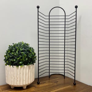 May include: A black metal plate rack with a curved top and base, featuring multiple horizontal bars for holding plates. A decorative, round, green plant sits in a white, textured pot with three small feet. The rack is approximately 15 inches tall.
