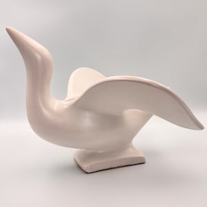 Peut inclure: Une sculpture en céramique rose pâle représentant un oiseau aux ailes déployées. La sculpture a une finition lisse et brillante et un design moderne et abstrait. La tête et le cou de l'oiseau sont allongés, et les ailes se courbent gracieusement vers l'extérieur. La base est de forme carrée.