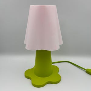 May include: A table lamp featuring a light pink, flared, petal-shaped shade atop a solid lime green base. The base is shaped like a five-petaled flower, and a matching green power cord with an inline switch extends from the side of the lamp.