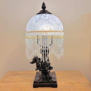 K&ouml;nnte beinhalten: Eine Tischlampe mit einem dunkelbraunen Sockel, der zwei Puttenfiguren zeigt. Die Lampe hat einen kuppelf&ouml;rmigen, wei&szlig; gesprenkelten Glasschirm mit einem dunklen Abschluss. Ein Behang aus klaren und blassgelben Glasperlen h&auml;ngt am Rand.