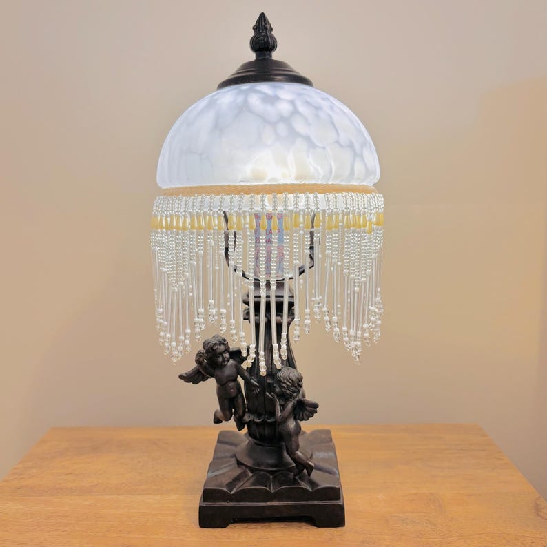 K&ouml;nnte beinhalten: Eine Tischlampe mit einem dunkelbraunen Sockel, der zwei Puttenfiguren zeigt. Die Lampe hat einen kuppelf&ouml;rmigen, wei&szlig; gesprenkelten Glasschirm mit einem dunklen Abschluss. Ein Behang aus klaren und blassgelben Glasperlen h&auml;ngt am Rand.