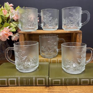 Peut inclure: Une tasse en céramique blanche avec une anse noire et l'inscription "But First, Coffee" en gras noir. La tasse est posée sur un fond flou, suggérant un cadre chaleureux.