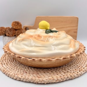 Puede incluir: Fuente de tarta de cerámica con una cobertura tipo merengue, con un acento de limón y hoja verde. La fuente de tarta es marrón con un borde con volantes, sobre un salvamanteles tejido. Una tabla de cortar de madera está en el fondo.