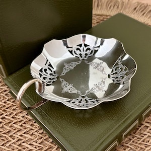 May include: A silver-coloured, scalloped-edge dish with an ornate handle. The dish features intricate, engraved floral patterns and openwork designs. The dish is likely made of metal and is displayed on a green surface.