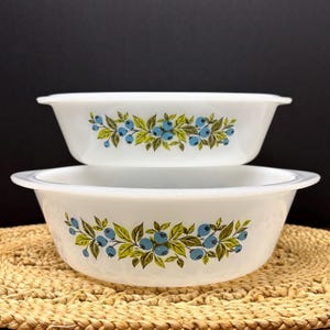 Puede incluir: Dos cuencos Pyrex vintage blancos de forma ovalada. Cada cuenco presenta un patrón decorativo de arándanos azules, hojas verdes y detalles amarillos. Los cuencos están apilados, destacando su diferencia de tamaño y diseño.