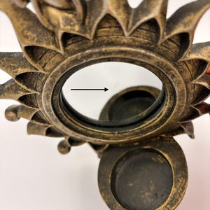 May include: A bronze-toned sun-shaped mirror with a distressed finish and a circular frame. A small black arrow points to a thin, faint scratch on the glass surface. The mirror is attached to a circular base with a recessed circular holder.