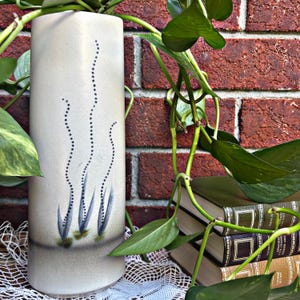 Peut inclure: Un vase en céramique blanc cassé de forme rectangulaire. Le vase présente un motif peint à la main de plantes aquatiques stylisées dans des tons de bleu et de vert. Le vase est entouré de vignes vertes et feuillues et se trouve devant un mur de briques.