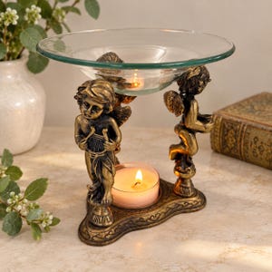 Puede incluir: Un quemador de aceite de metal dorado con dos figuras de querubines que sostienen un cuenco de cristal transparente. La base es triangular con detalles ornamentales y una vela de té encendida se sitúa entre los querubines para calentar.