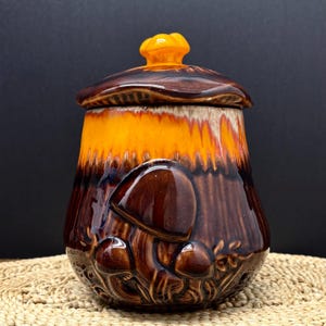 May include: A ceramic mushroom-shaped jar with a lid. The jar is glazed in shades of brown and orange, with a textured mushroom design on the front. The lid has a small, yellow mushroom-shaped handle.