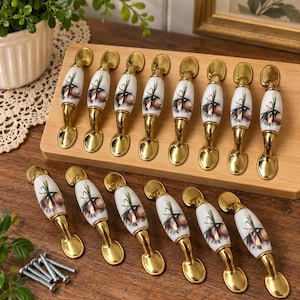 May include: A set of fourteen cabinet pulls featuring a white ceramic center with a painted onion and green sprout design. Each pull has polished gold-tone metal ends and a curved handle shape. A small pile of metal mounting screws is included.