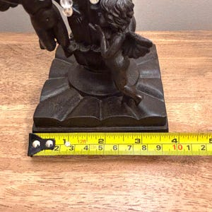 K&ouml;nnte beinhalten: Eine dunkelbraune Metallskulptur mit zwei Puttenfiguren, die um einen zentralen Sockel auf einer quadratischen Basis angeordnet sind. Ein gelbes Ma&szlig;band liegt davor und zeigt, dass die Basis etwa 10 cm breit ist.
