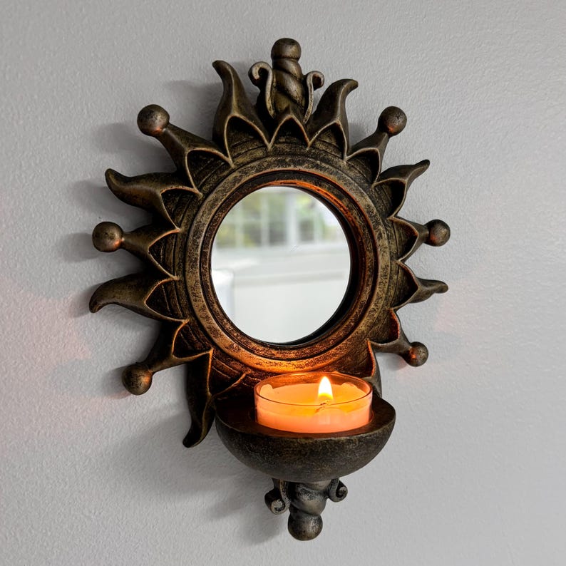 May include: A wall-mounted candle holder featuring a circular mirror surrounded by a dark bronze-toned sunburst frame with pointed rays. A small, semi-circular shelf at the base holds a glass votive cup containing a lit orange candle.