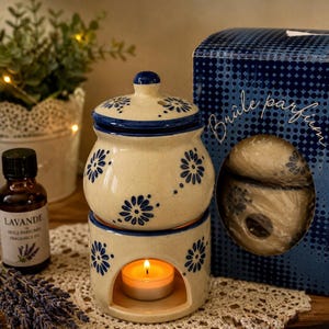 May include: A cream-colored ceramic oil burner with a lid and blue floral patterns. The base features an arched opening containing a lit tea light candle. Beside it sits a bottle labeled Lavande and a blue box with the text Brûle parfum.