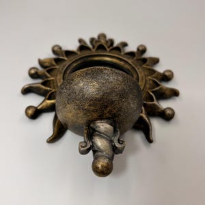 May include: A wall-mounted basin with a sunburst design featuring a circular center and radiating rays. The object has a distressed bronze finish with dark highlights and a twisted, rope-like decorative element extending downward.