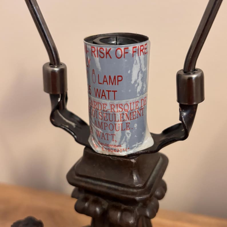 K&ouml;nnte beinhalten: Eine dunkelbraune Lampenfassung aus Metall mit einem zylindrischen Warnetikett, das roten Text mit der Aufschrift RISK OF FIRE, LAMP, 25 WATT sowie franz&ouml;sischen Text enth&auml;lt. Die Fassung wird von einem Metallrahmen mit zwei Armen gehalten.
