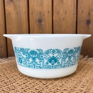 May include: White Pyrex bowl with teal floral design. The bowl has a rounded shape with two handles and a decorative band of teal floral patterns around the exterior. The bowl is likely vintage and suitable for kitchen use.