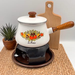 Op de afbeelding: Witte emaille fonduepan met een houten handvat en deksel. De pan heeft een kleurrijk fruit- en groentendesign met de tekst "La Fondue". Hij staat op een zwarte standaard met een bruine basis. Een houten snijplank staat op de achtergrond.