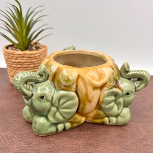 May include: A ceramic planter featuring two light green elephant figures on the sides with a central bowl finished in a glossy tan and brown glaze. The elephants have raised trunks and detailed ears, forming a decorative base for the central opening.