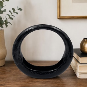 Peut inclure: Un vase creux, noir et circulaire, doté d'une finition lisse et brillante. L'objet présente un large centre ouvert et une base plate, créant une silhouette en forme d'anneau. La surface reflète la lumière, soulignant la couleur sombre et uniforme.